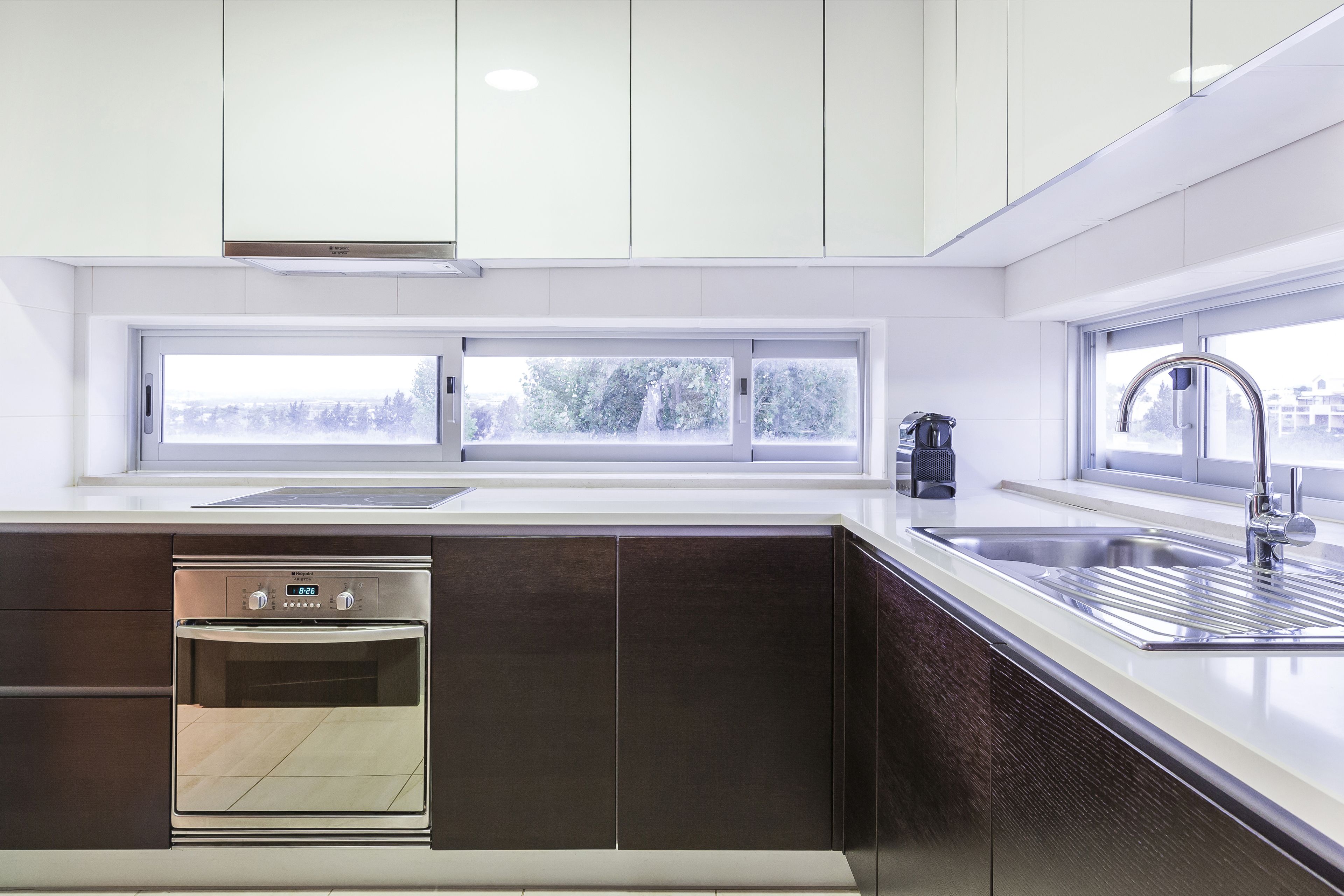 A modern kitchen with white cabinets, a built-in oven, a stainless steel sink, and a window with a view of trees.
