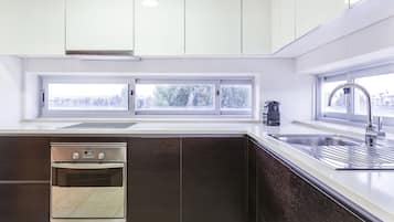 A modern kitchen with white cabinets, a built-in oven, a stainless steel sink, and a window with a view of trees.