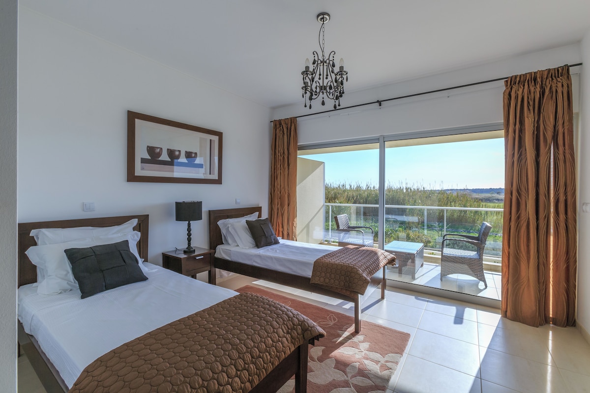 A hotel room with two beds, a chandelier, a balcony with a table and chairs, and a view of the landscape.