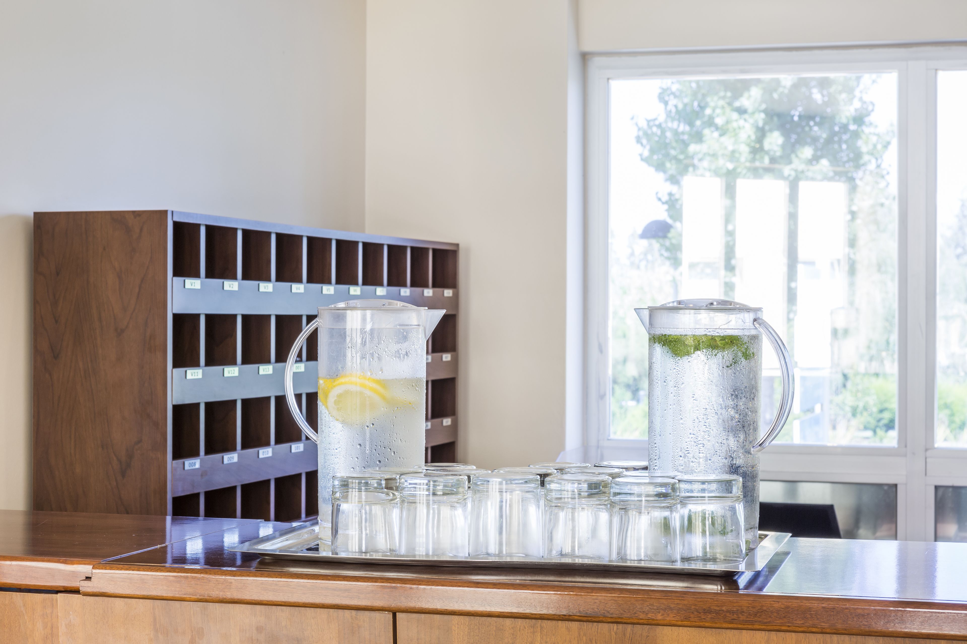 A kitchen counter with a water dispenser, a pitcher with a lemon slice, and a row of glasses.