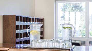 A kitchen counter with a water dispenser, a pitcher with a lemon slice, and a row of glasses.