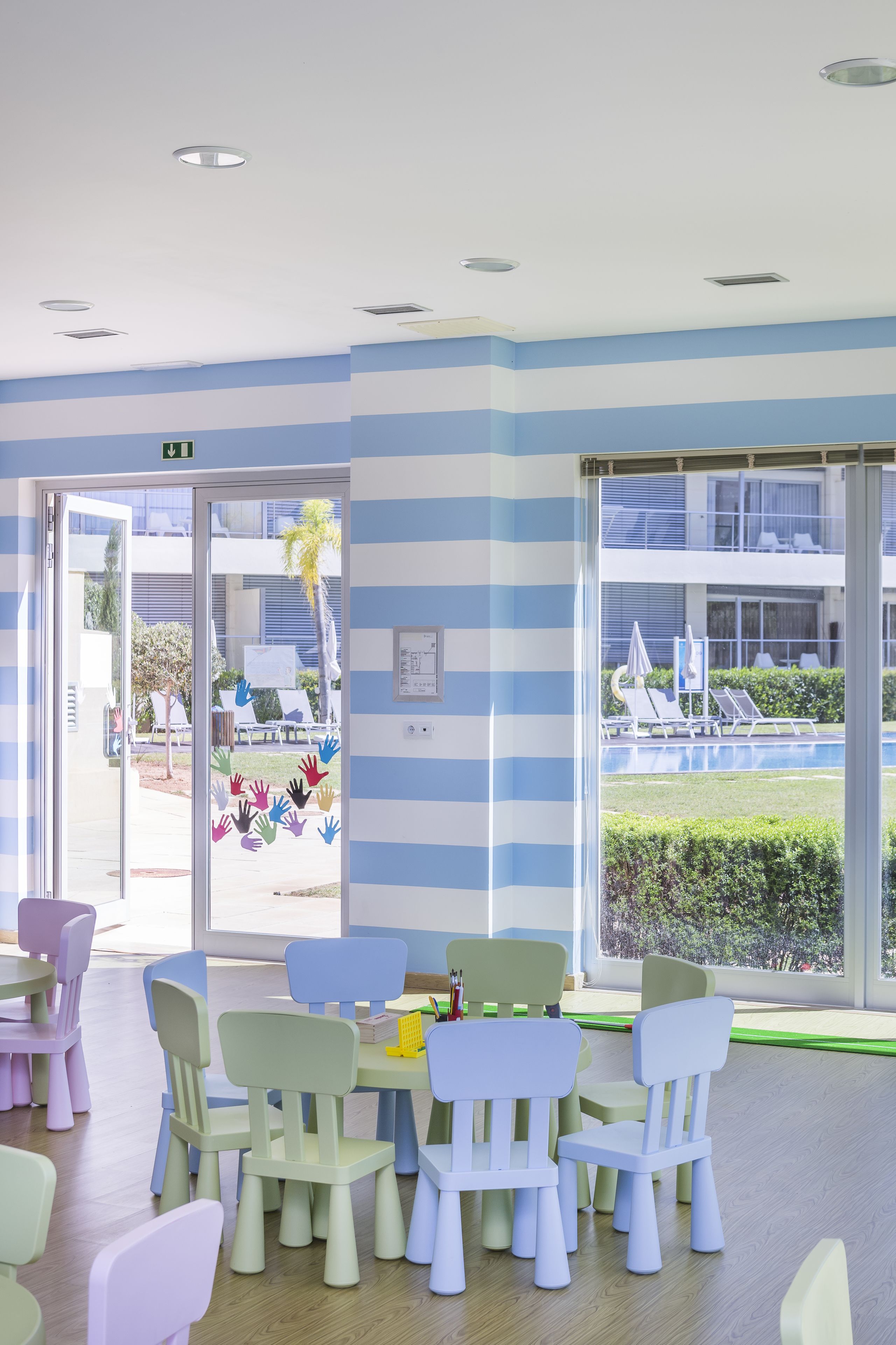 A brightly lit room with striped walls, a large window with a view of a pool and outdoor seating, and several tables with chairs.