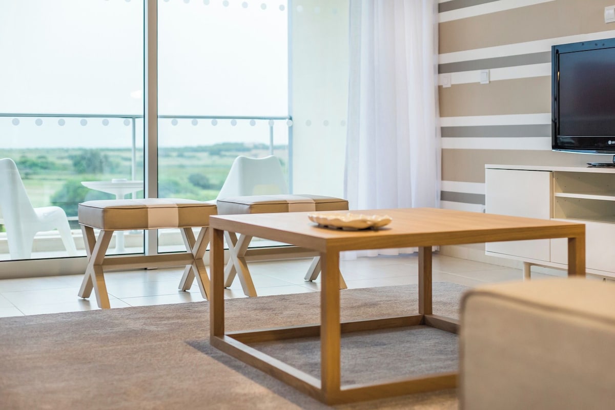 A modern living room with a wooden table, two chairs, a television, and a view of the outdoors.