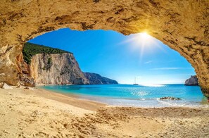 A beach with a natural arch formation, clear blue water, and a distant sailboat. - Village Villas (Lefkada)