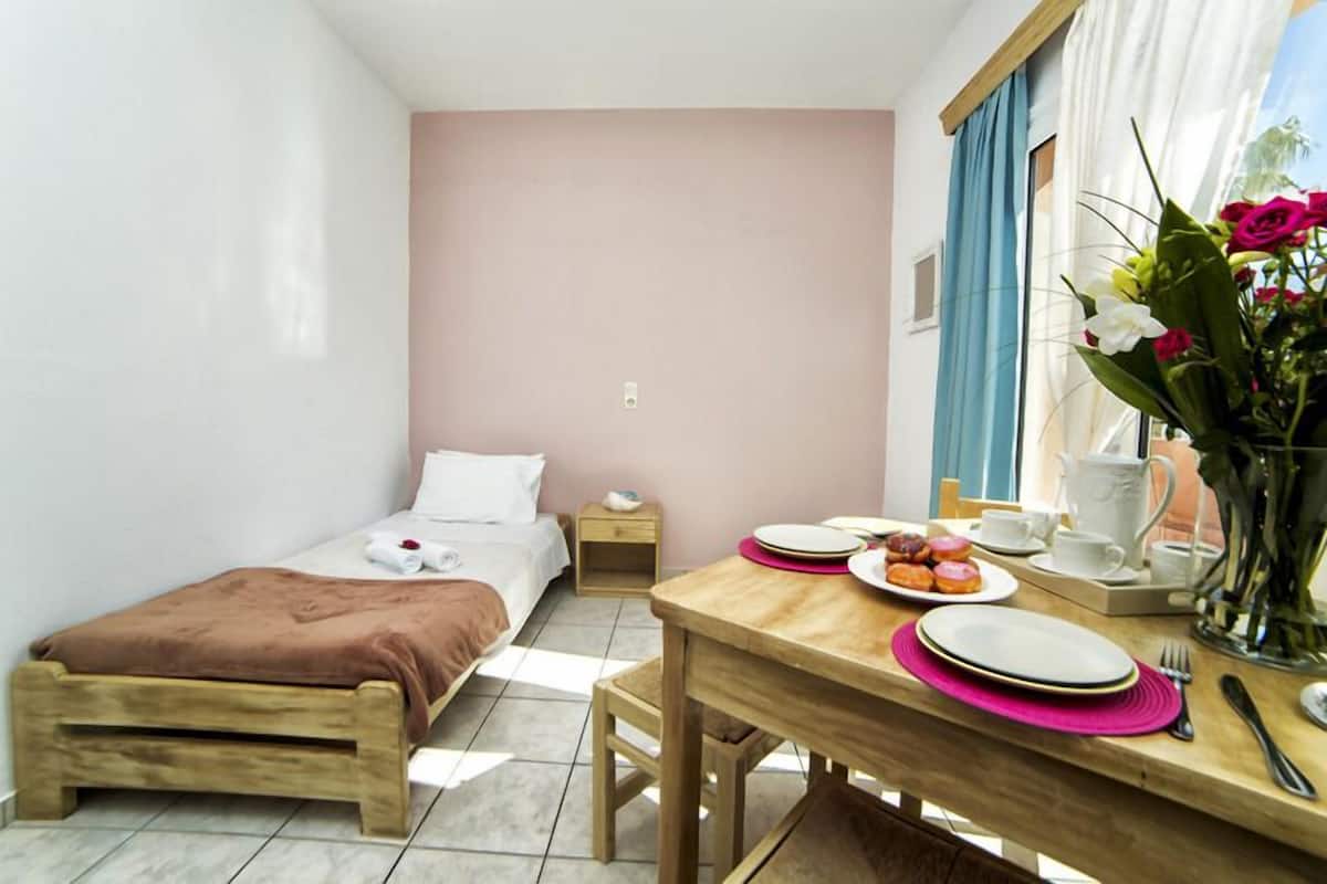 A small, well-lit room with a single bed, a wooden table with breakfast items, and a window with curtains.