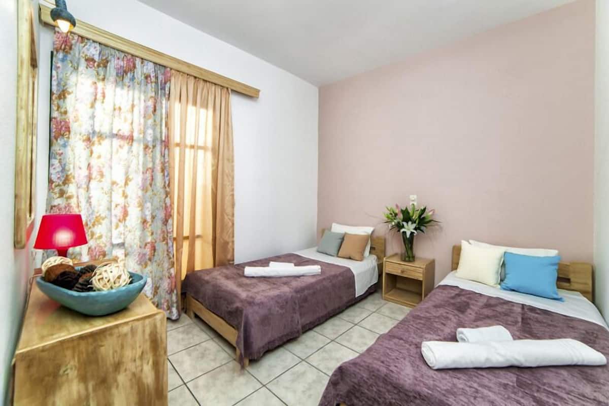 A hotel room with two beds, a nightstand, a lamp, and a window with curtains.