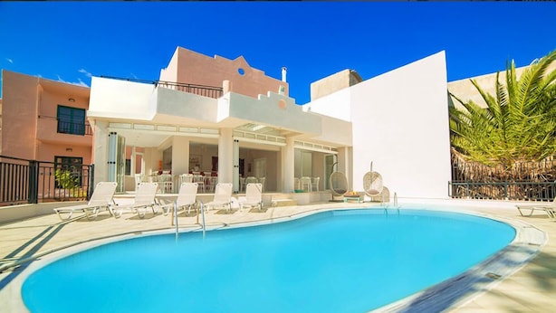 A swimming pool with lounge chairs, a covered seating area, and a building with a flat roof and balconies. - Malia Central (Hersonissos)