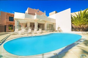 A swimming pool with lounge chairs, a covered seating area, and a building with a flat roof and balconies. - Malia Central (Hersonissos)