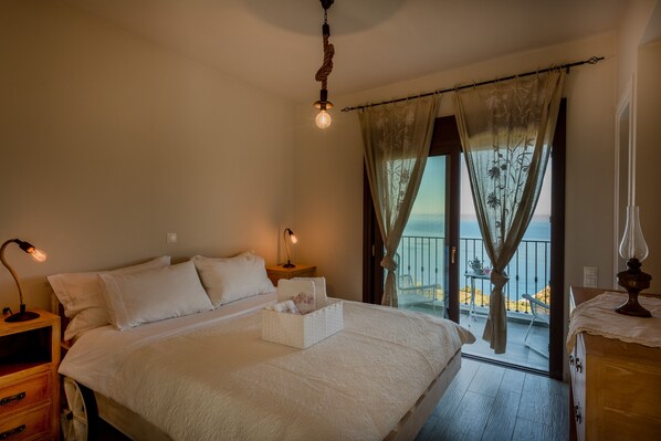 A bedroom with a large bed, a bedside table with a lamp, a nightstand, and a view of the sea through a balcony.