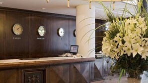 A reception area with a dark wood reception desk, a round table with a vase of flowers, and a chandelier with multiple light fixtures. - Dos Reyes Hotel (Mar del Plata)