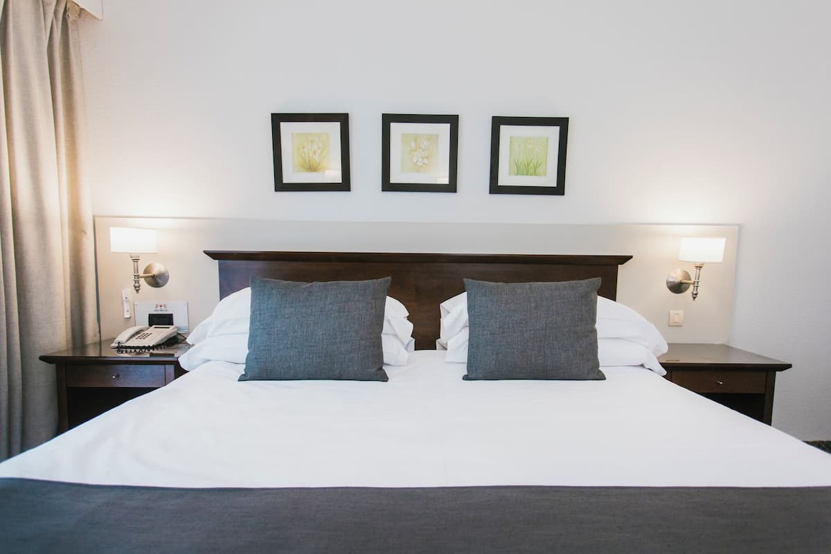 Una cama bien hecha con sábanas blancas y dos almohadas grises, flanqueada por dos mesitas de noche con lámparas y un teléfono. Tres cuadros enmarcados adornan la pared sobre la cama.