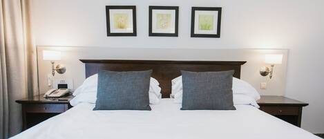 A neatly made bed with white linens and two grey pillows, flanked by two bedside tables with lamps and a phone. Three framed artworks are hung on the wall above the bed.