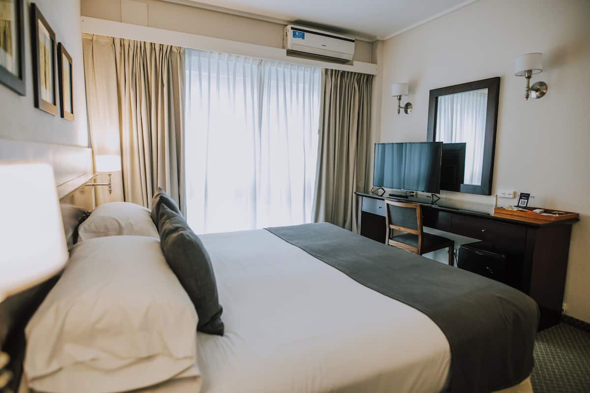 Habitación de hotel con cama, escritorio, silla, televisor y espejo.