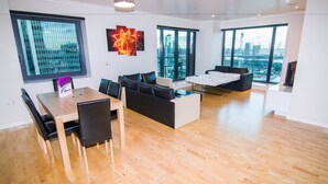 A spacious living area with a wooden floor, a dining table with chairs, a black sofa, a white coffee table, and large windows offering views of the city. - Canary Wharf - Corporate Riverside Apartments (London)