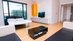 A modern living room with a sofa, a coffee table, a TV, and a large window offering a city view. - Canary Wharf - Corporate Riverside Apartments (London)
