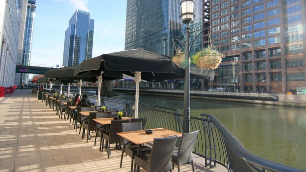 Outdoor seating with tables and chairs, a canal, and modern buildings in the background. - Canary Wharf - Corporate Riverside Apartments (London)