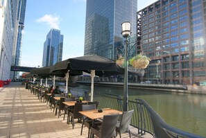 Terrasse mit Tischen und Stühlen, ein Kanal und moderne Gebäude im Hintergrund.