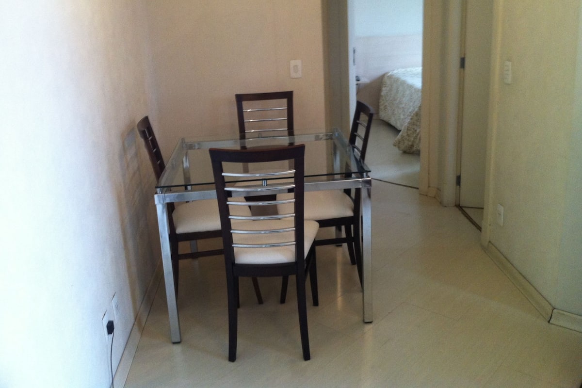A dining area with a glass-top table and four chairs, a light-colored floor, and a plain wall.
