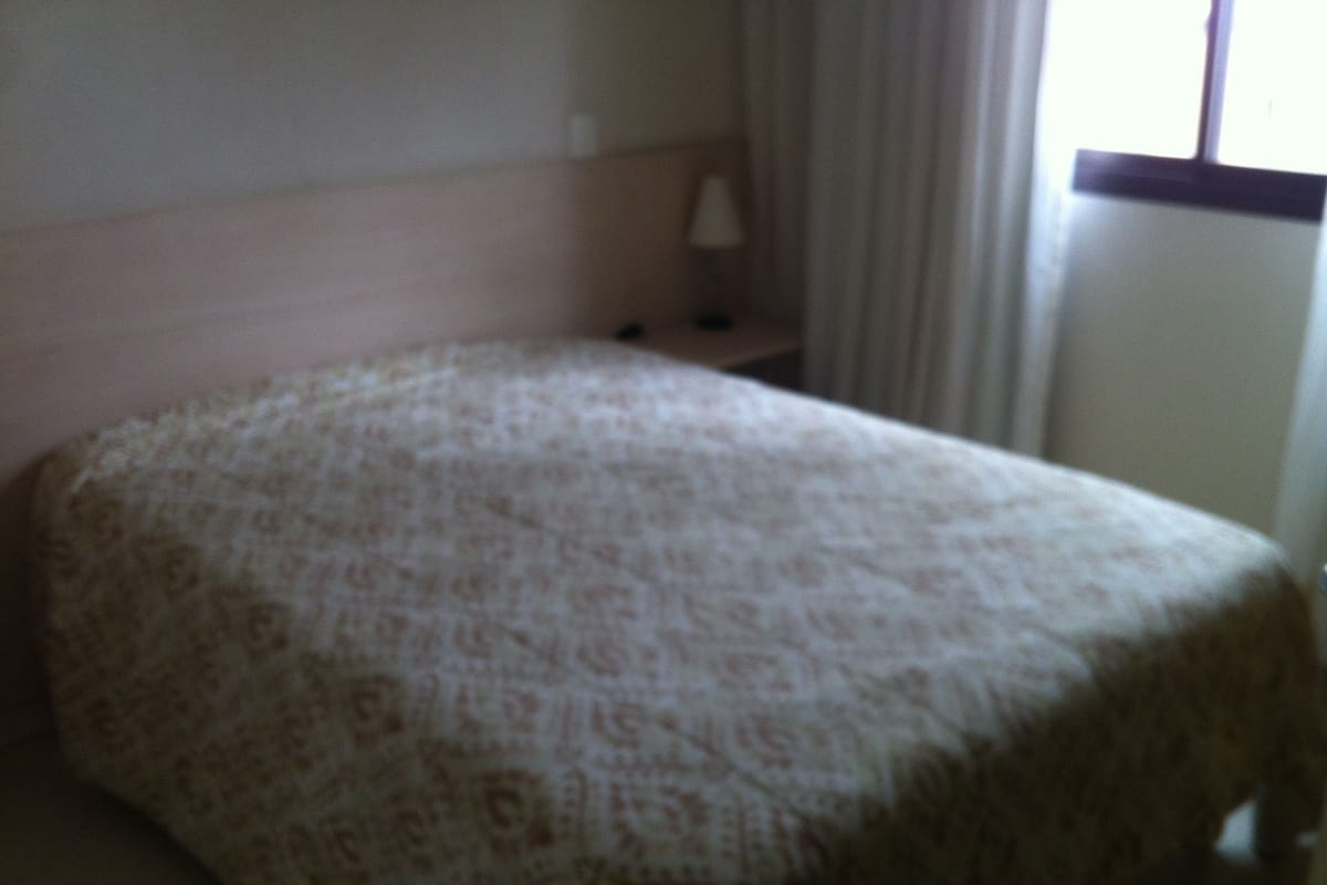 A small, neatly made bed with a patterned bedspread in a simple room with a nightstand and a lamp.