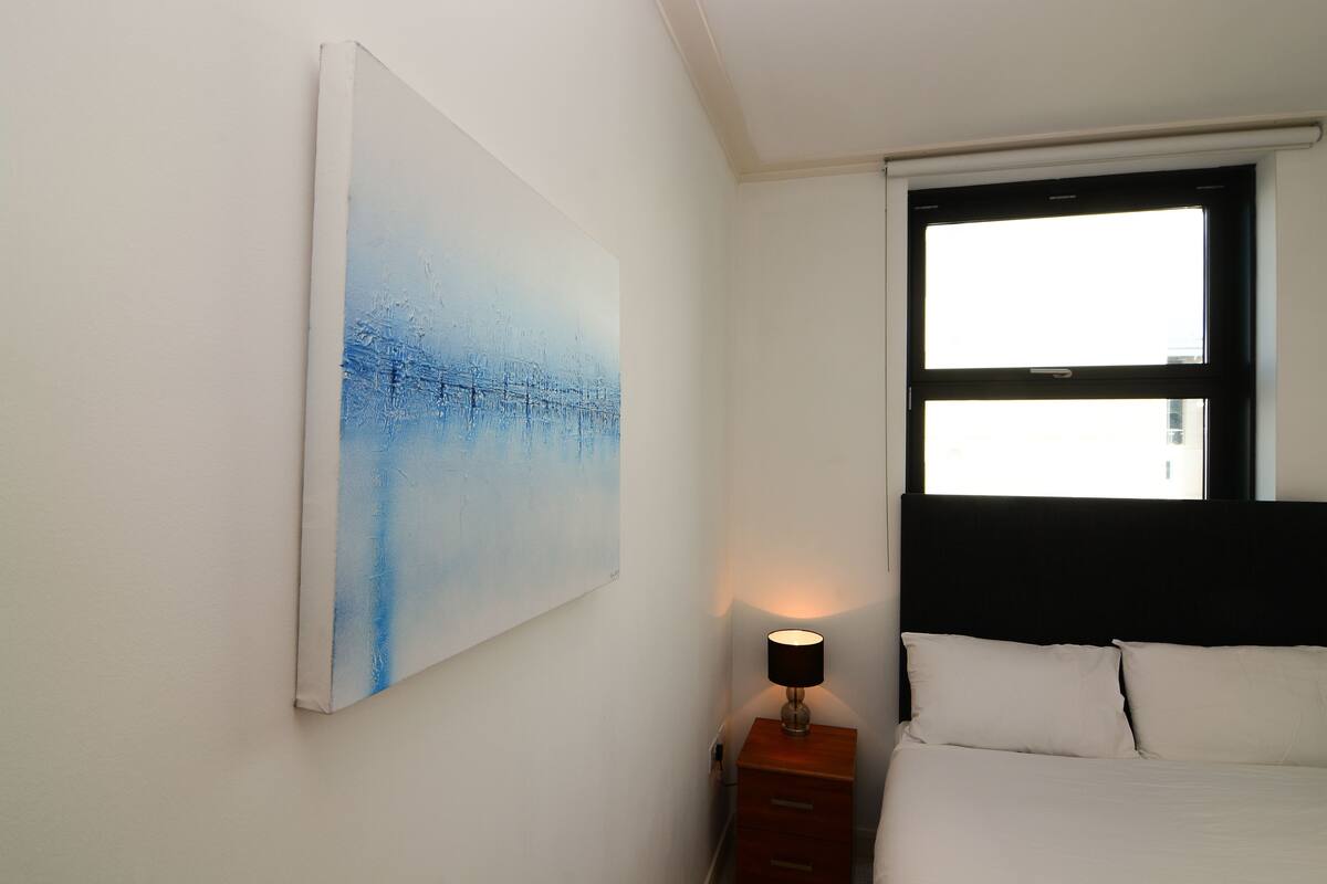 Un dormitorio con cama, mesita de noche, lámpara y un cuadro abstracto grande en la pared.