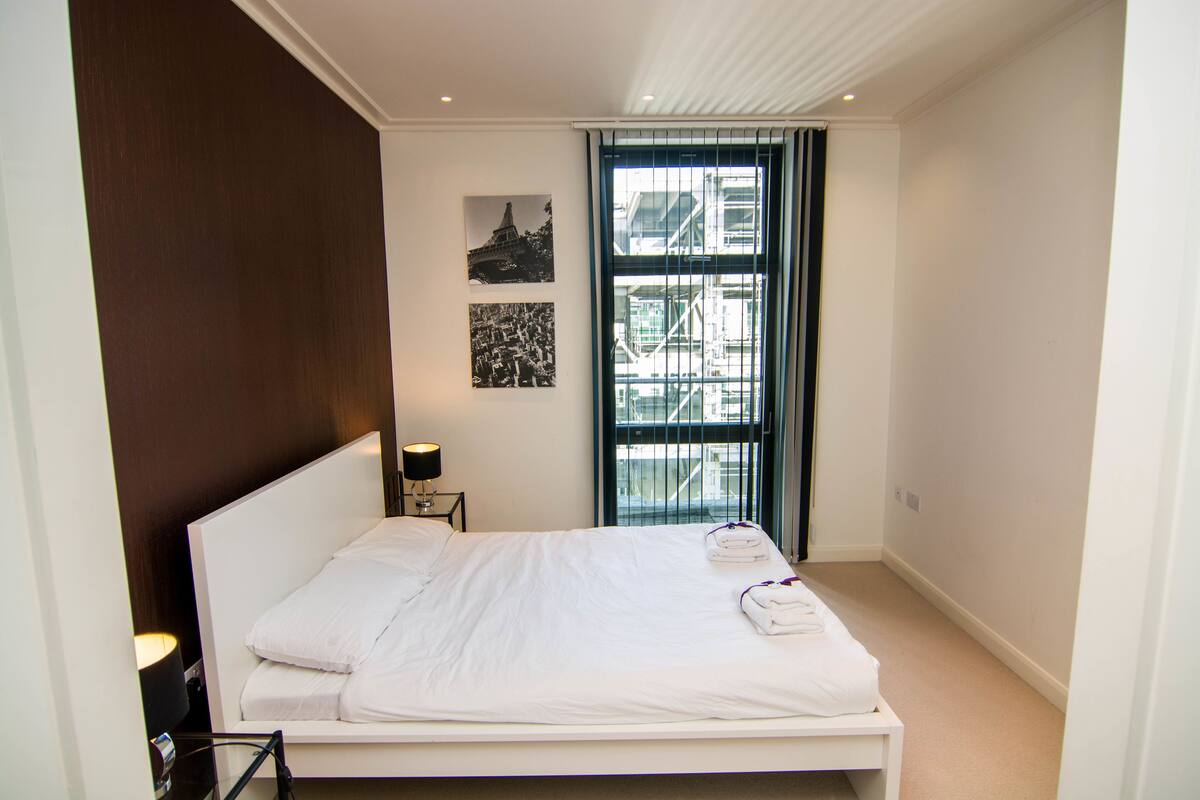 Una habitación de hotel con una cama grande, una mesita de noche con una lámpara y una ventana que ofrece vistas a un edificio.