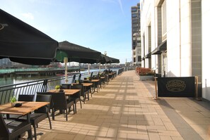 Een terras aan de rivier met tafels en stoelen, een overdeeld gedeelte en een banner die een vrijhuis aangeeft.