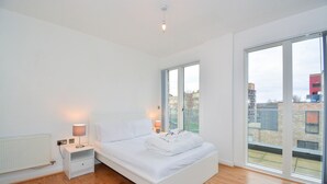 Quarto moderno com uma cama grande, mesas de cabeceira brancas e vista para edifícios através de uma janela ampla.