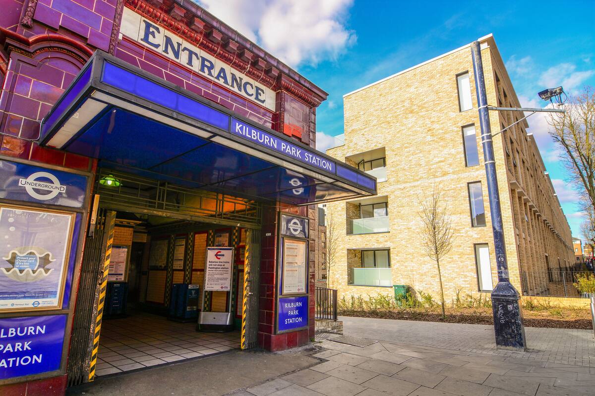 The entrance to Kilburn Park Station with a clear sign, a tiled floor, and a modern building adjacent to it.