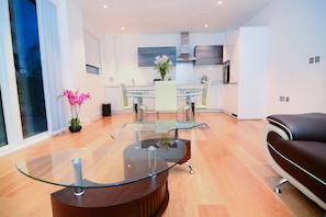 A modern living room with a glass coffee table, a leather sofa, and a dining area with a glass table and chairs. - Apartment Wharf - Cambridge Avenue (London)