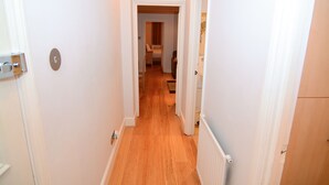 Un pasillo estrecho con suelo de madera y paredes blancas, que conduce a una habitación con cama y sillón.