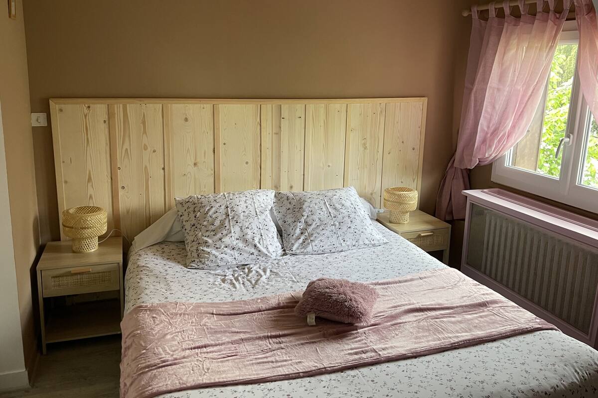 A bedroom with a wooden headboard, a bed with floral bedding, a pink throw blanket, and a window with curtains.