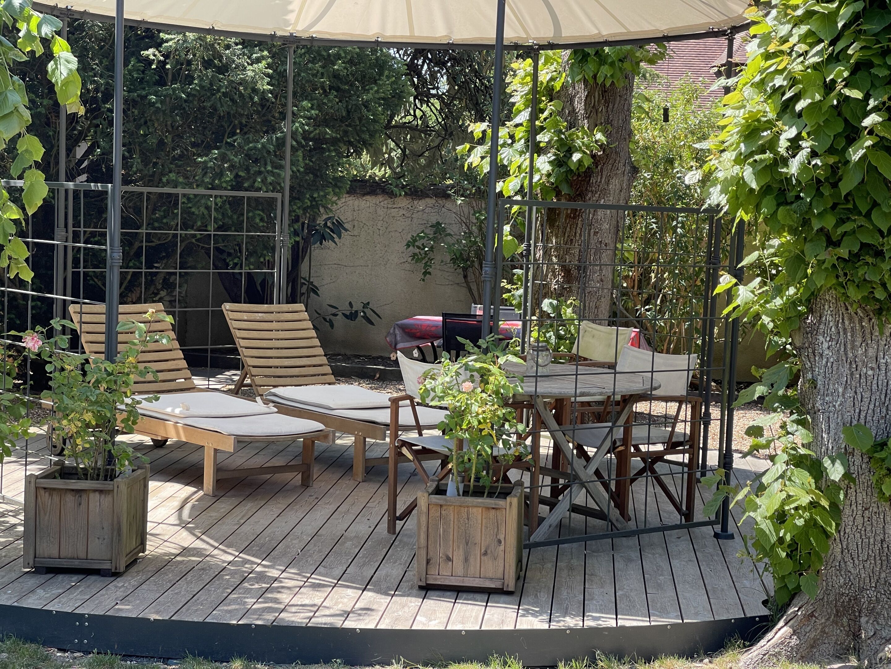 An outdoor patio with wooden furniture, including lounge chairs and a table, surrounded by greenery and a pergola.