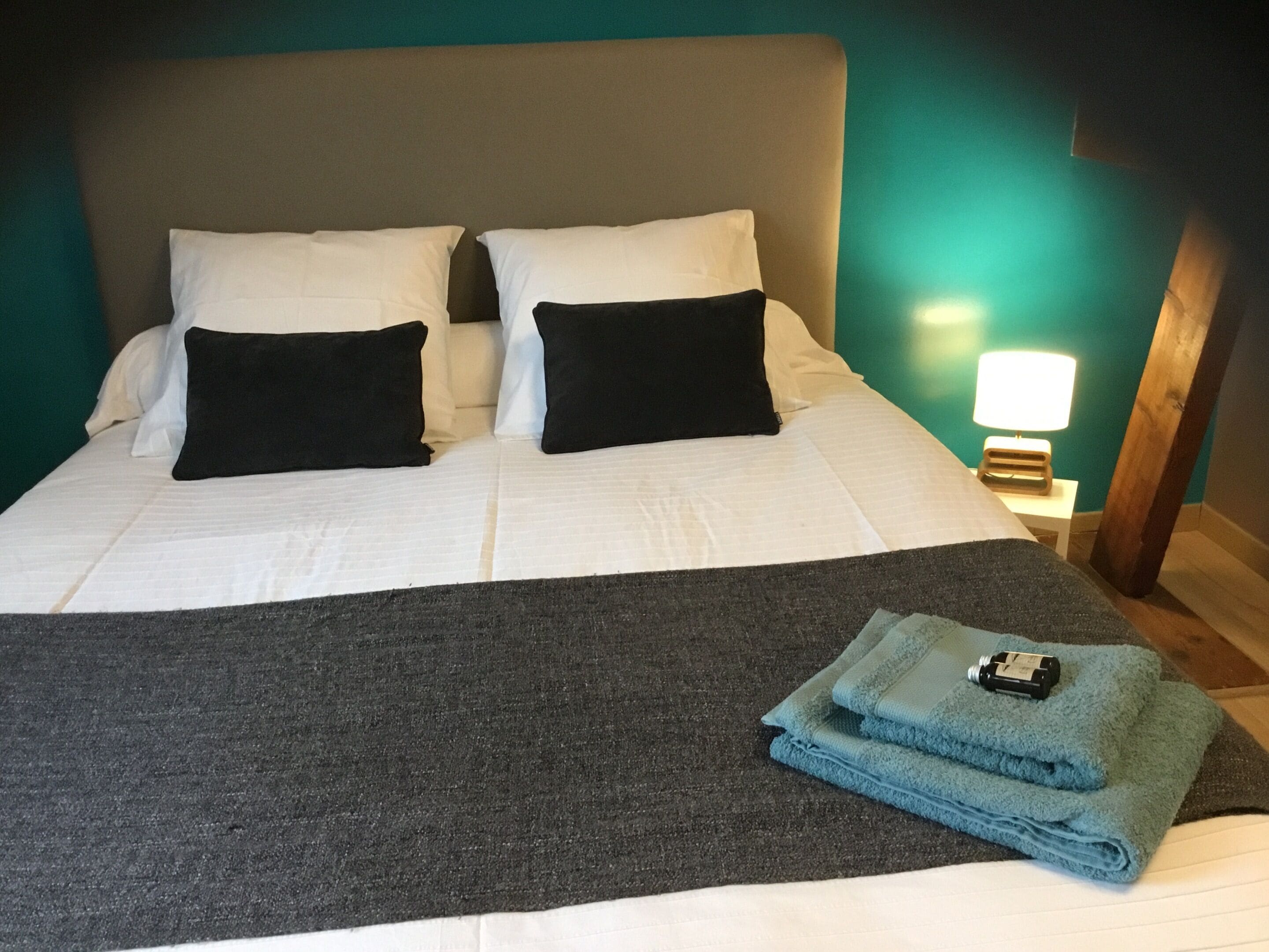Una cama bien hecha con almohadas blancas y negras, una manta gris y una toalla azul doblada con una botella encima.