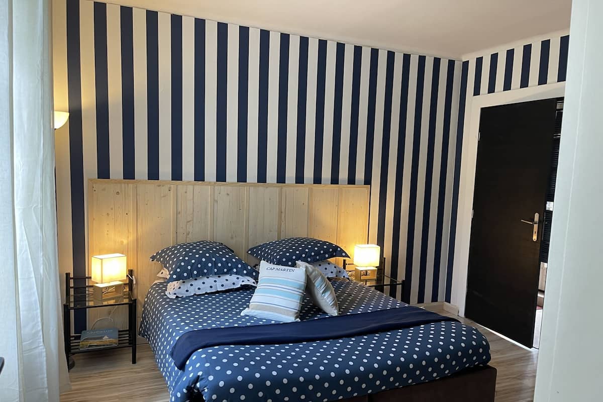 A bedroom with a striped wall, a wooden headboard, two bedside tables with lamps, a bed with polka dot bedding, and a door to the right.