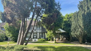A traditional Tudor-style house with a well-maintained garden, a patio with outdoor furniture, and a large tree in the foreground. - La Collinière (Sainte-Gemme-Moronval)
