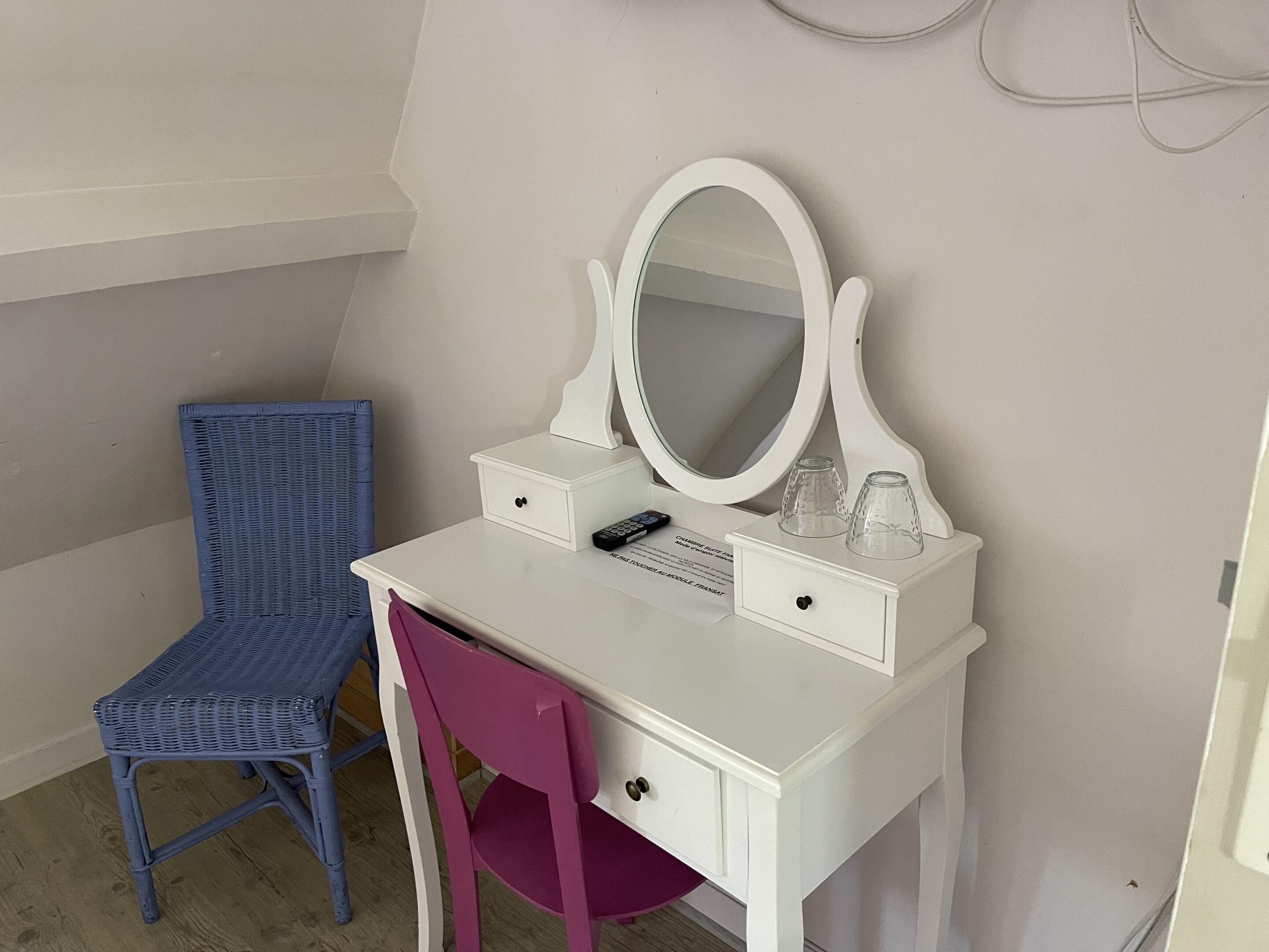 A white vanity with a large mirror, two drawers, and a small table with a remote control and glasses. A blue chair and a pink chair are positioned in front of the vanity.