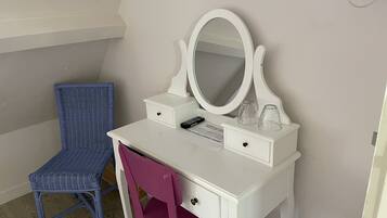 A white vanity with a large mirror, two drawers, and a small table with a remote control and glasses. A blue chair and a pink chair are positioned in front of the vanity.
