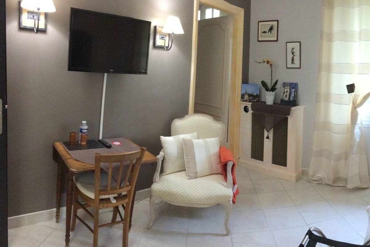 A room with a TV mounted on the wall, a dining table with chairs, a sofa, a small table with a lamp, and a window with curtains.