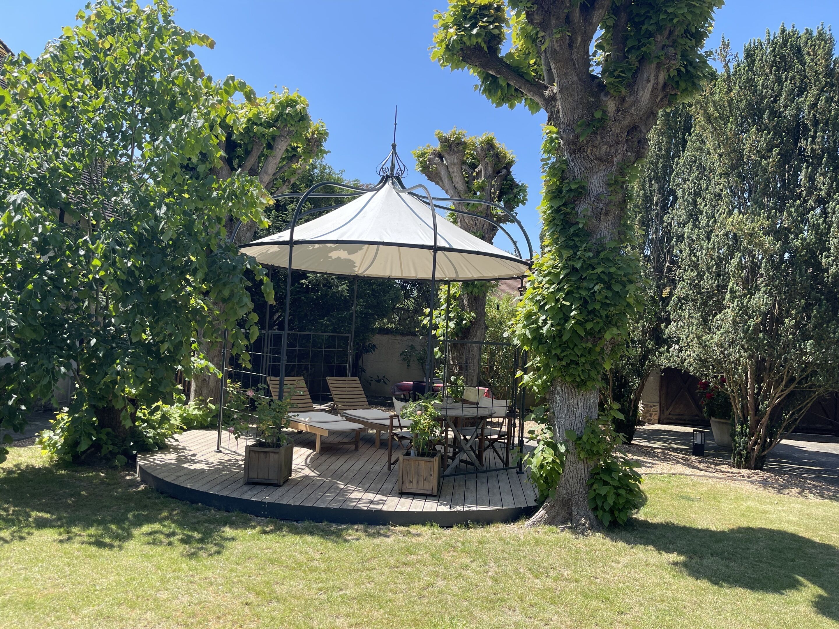 A covered outdoor seating area with a table and chairs, surrounded by trees and a pergola.