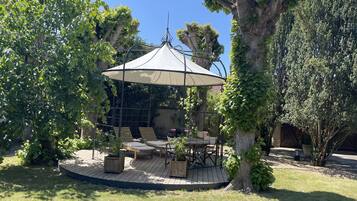 A covered outdoor seating area with a table and chairs, surrounded by trees and a pergola.