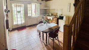 A kitchen with wooden beams, a dining table with chairs, a radiator, and a window with curtains. - La Collinière (Sainte-Gemme-Moronval)
