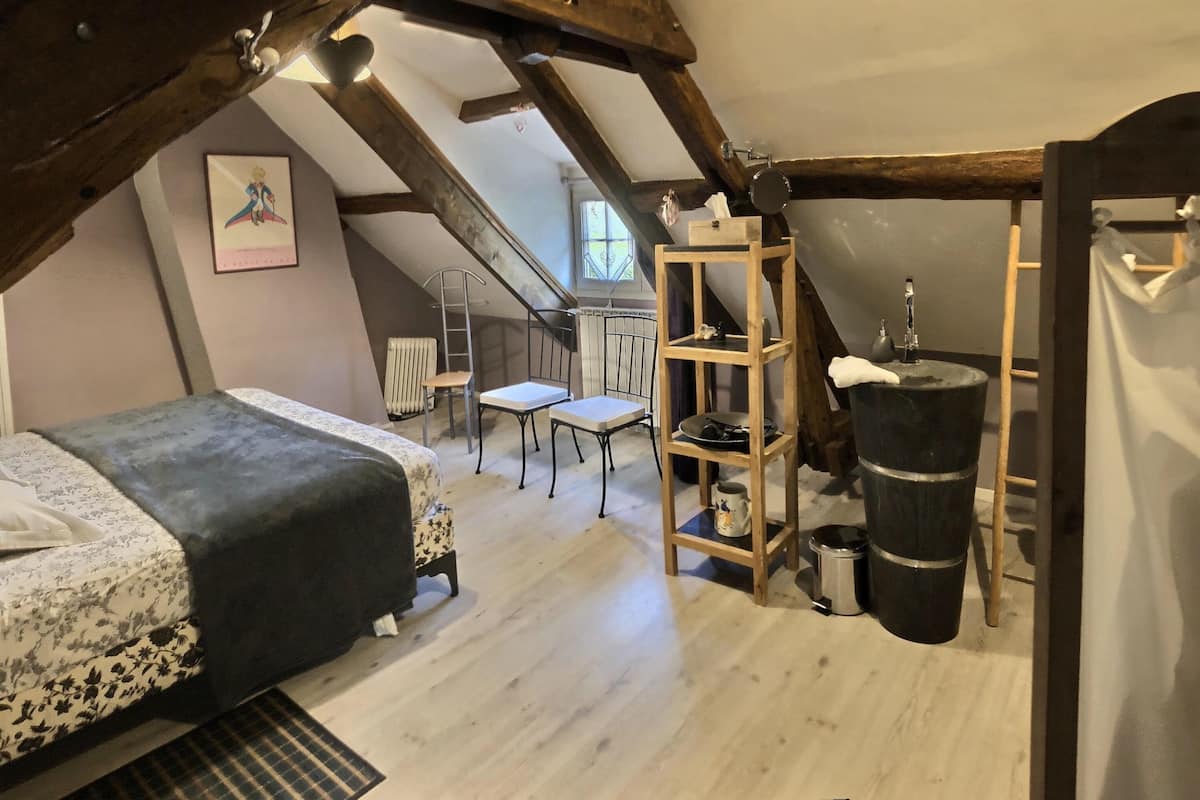 A cozy attic bedroom with a bed, a small table, a chair, a radiator, and a wooden shelf with a basin.