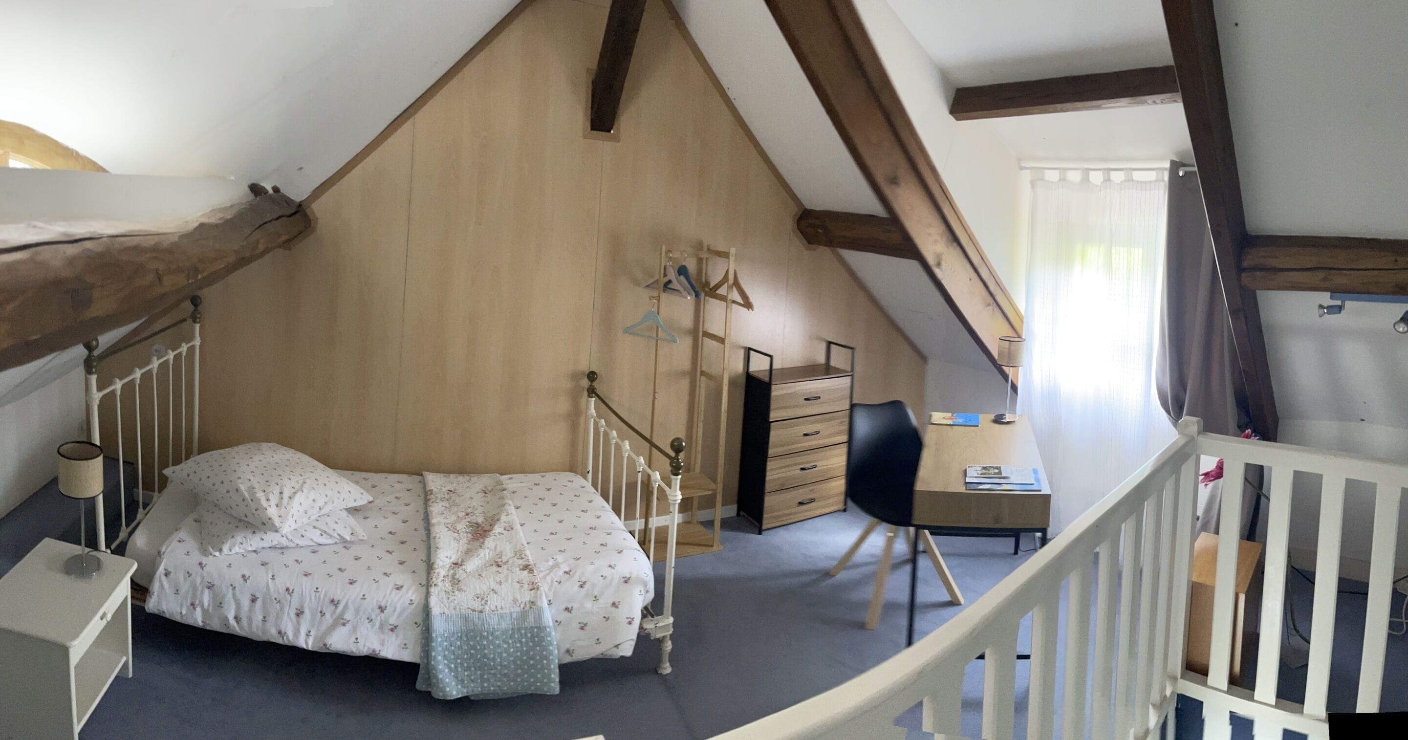 Une chambre avec un plafond en pente, un lit recouvert d’une couette à motifs floraux, une commode en bois, un bureau avec une chaise et une rambarde blanche.