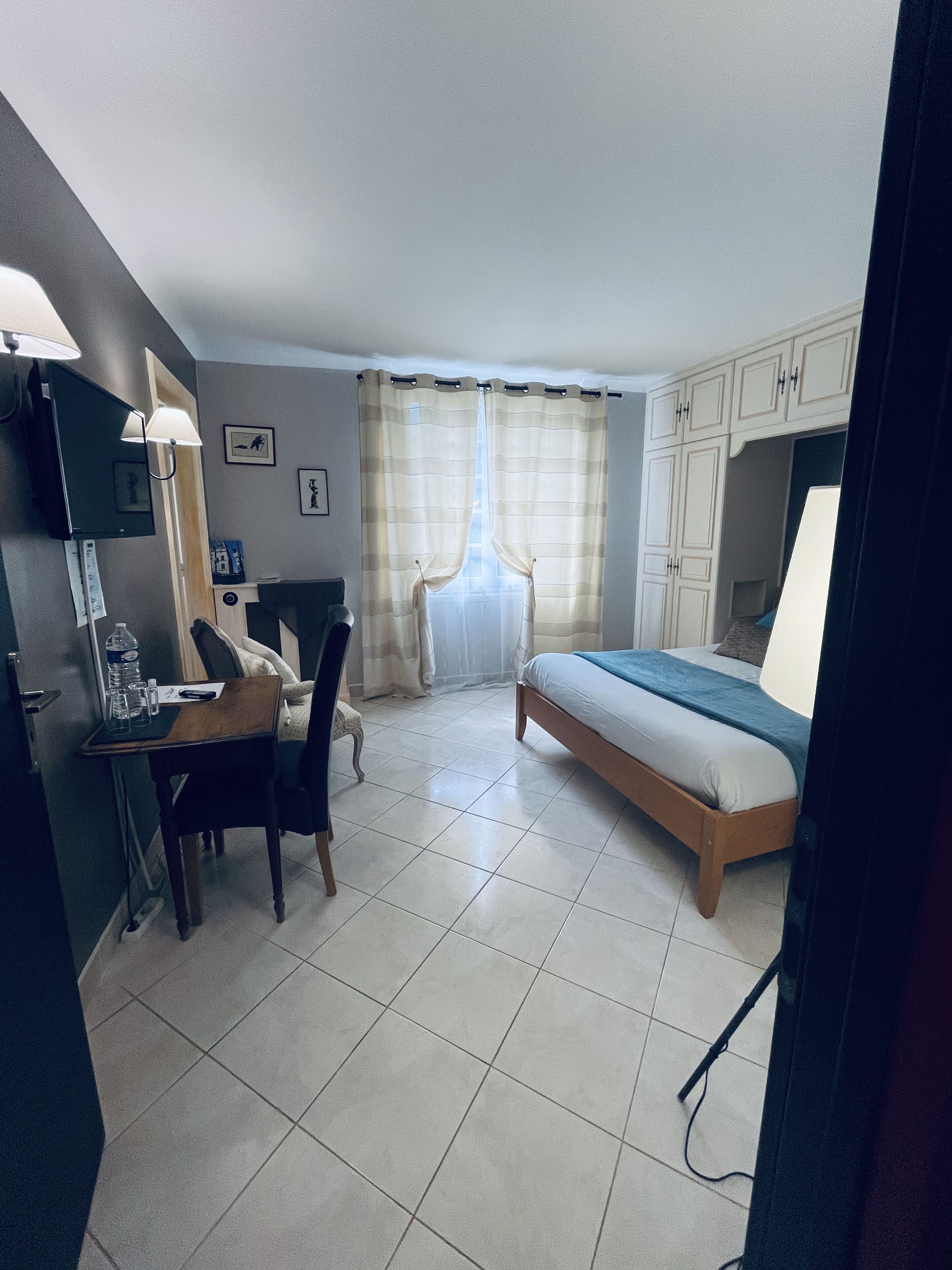 A bedroom with a bed, a desk, a chair, a TV, and a window with curtains.