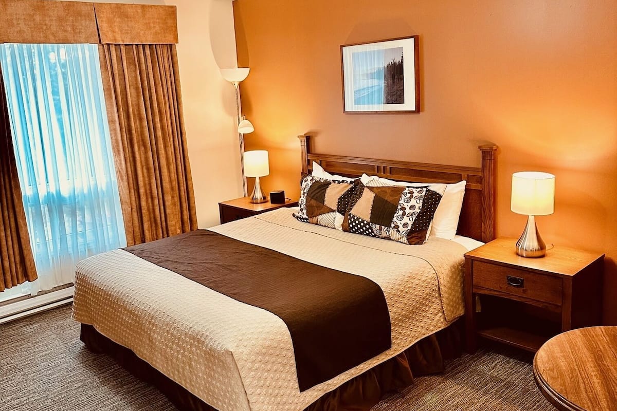 A hotel room with a large bed, two bedside tables with lamps, a round table, a chair, a ceiling light fixture, a window with curtains, and a framed picture on the wall.