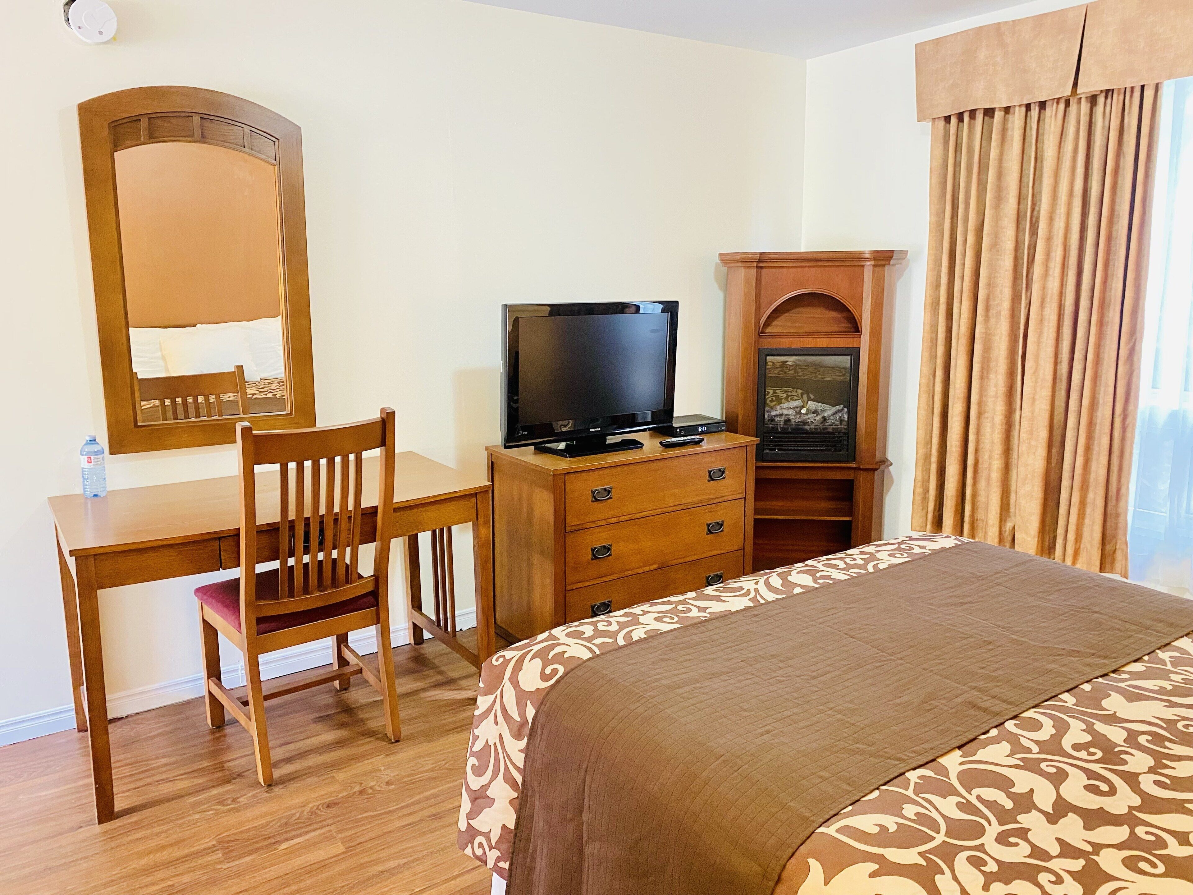 Habitación de hotel con un escritorio de madera, una silla, un televisor sobre un mueble de madera, una cama con colcha estampada y un espejo grande.