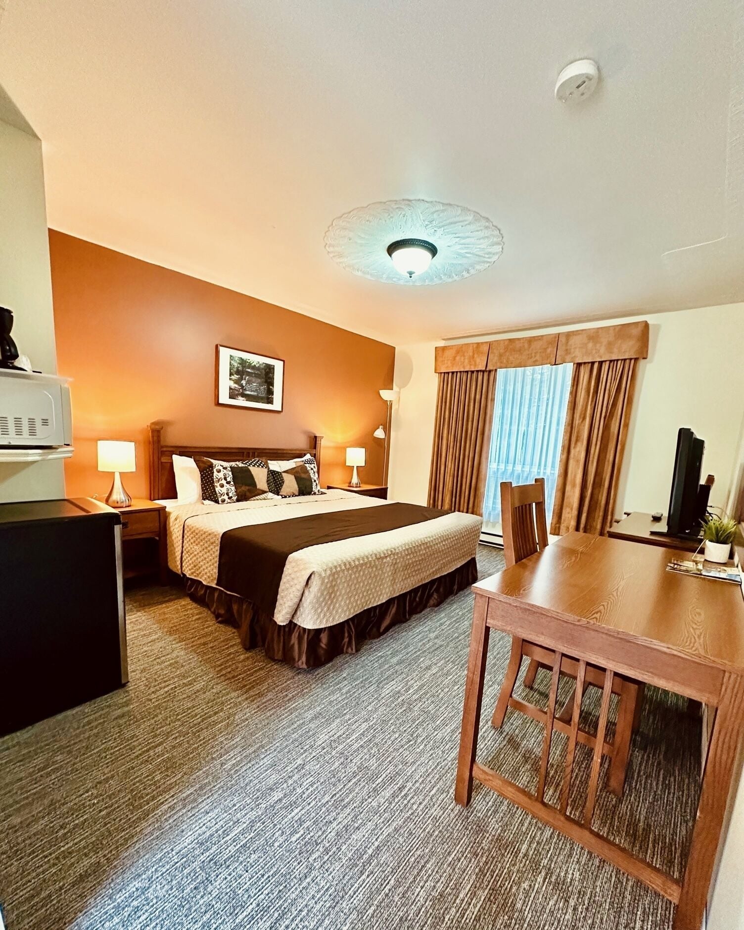 A hotel room with a bed, bedside tables, a desk, a chair, a television, and a window with curtains.