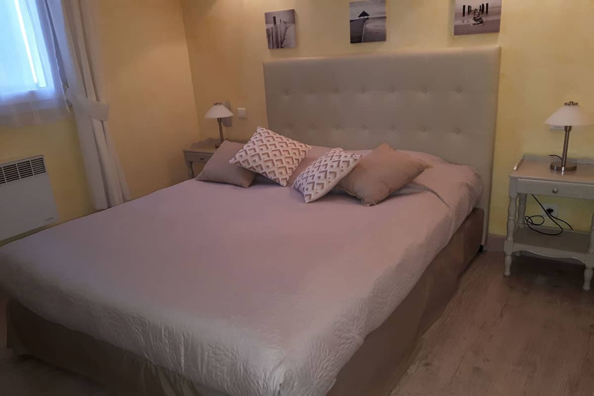 A bedroom with a large bed, two bedside tables, a wall-mounted lamp, and a window with curtains.