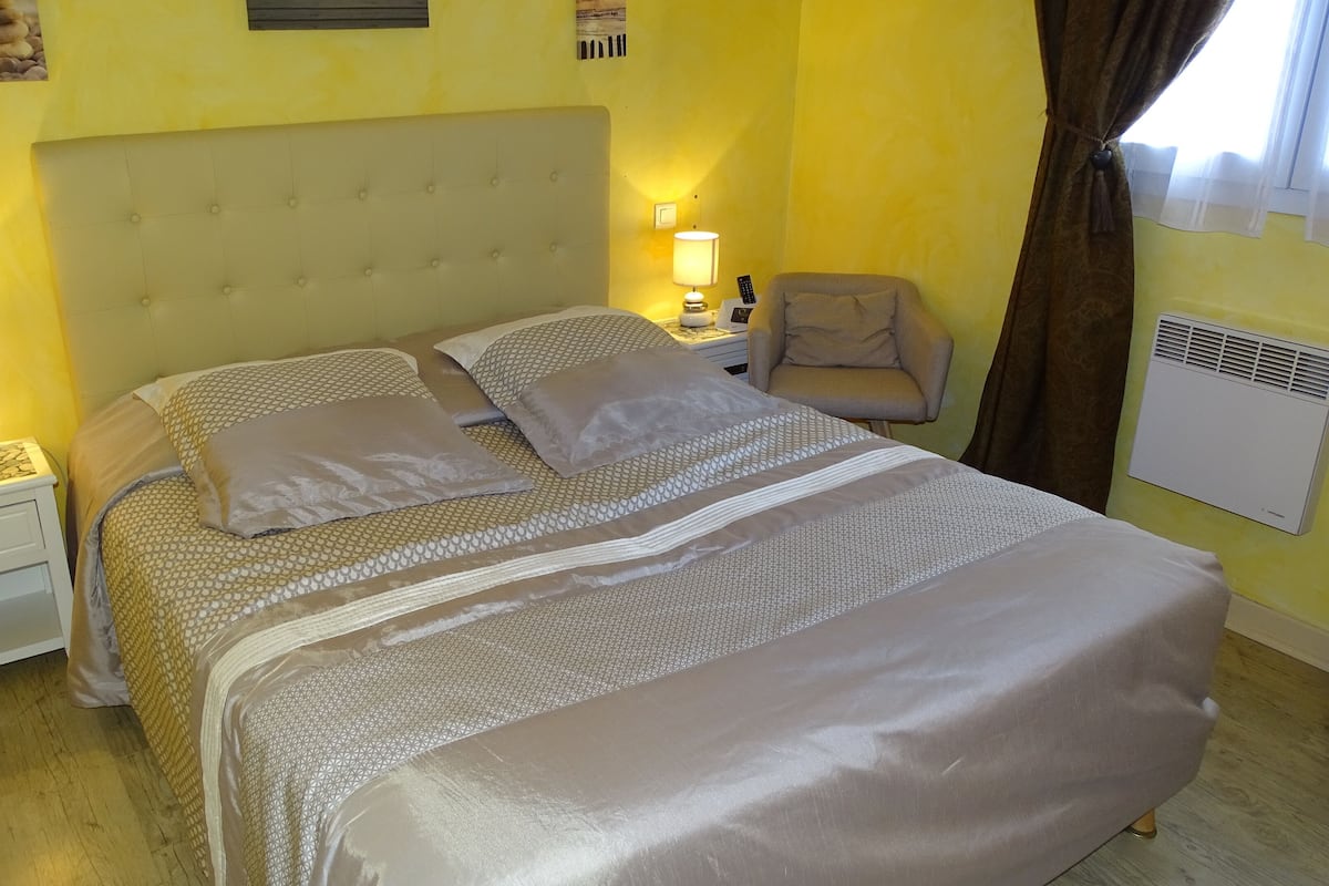 A neatly made bed with a patterned bedspread and pillows, a bedside lamp, a chair, and a wall-mounted air conditioner.