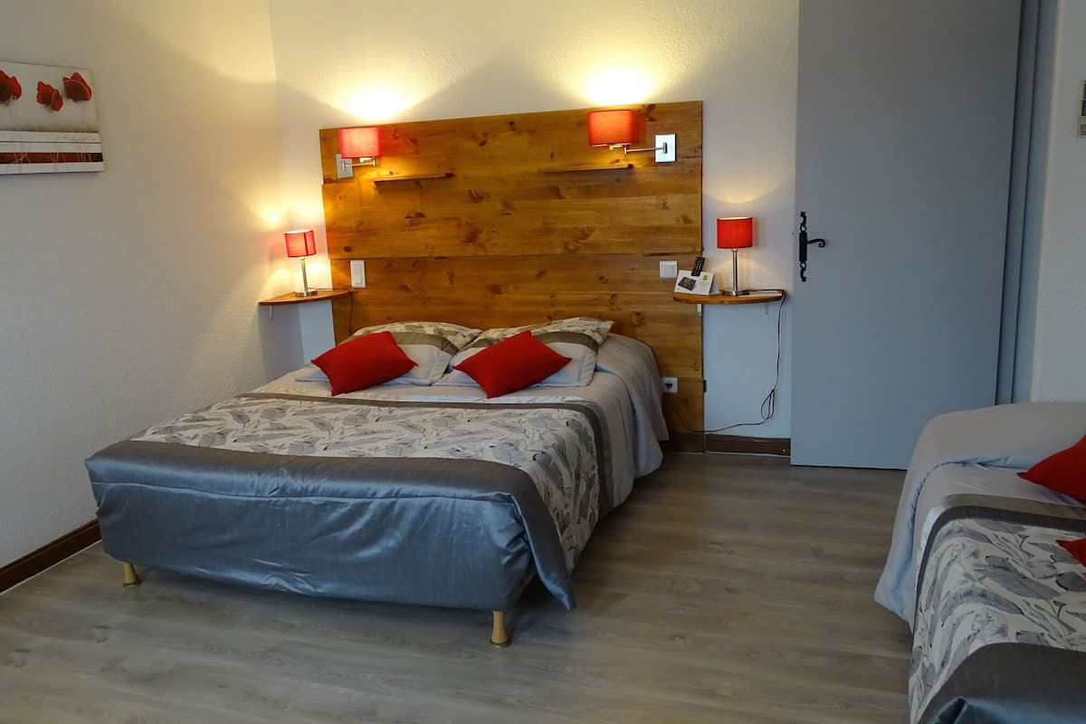 A bedroom with a wooden headboard, two bedside tables with lamps, a bed with a grey bedspread and red pillows, and a small wooden shelf.